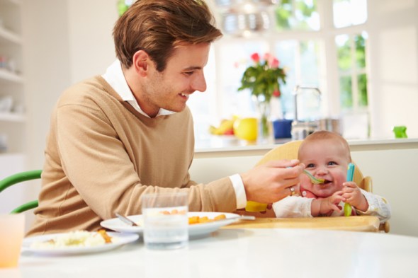 Father Feeding Baby Sitting In High Chair At Mealtime